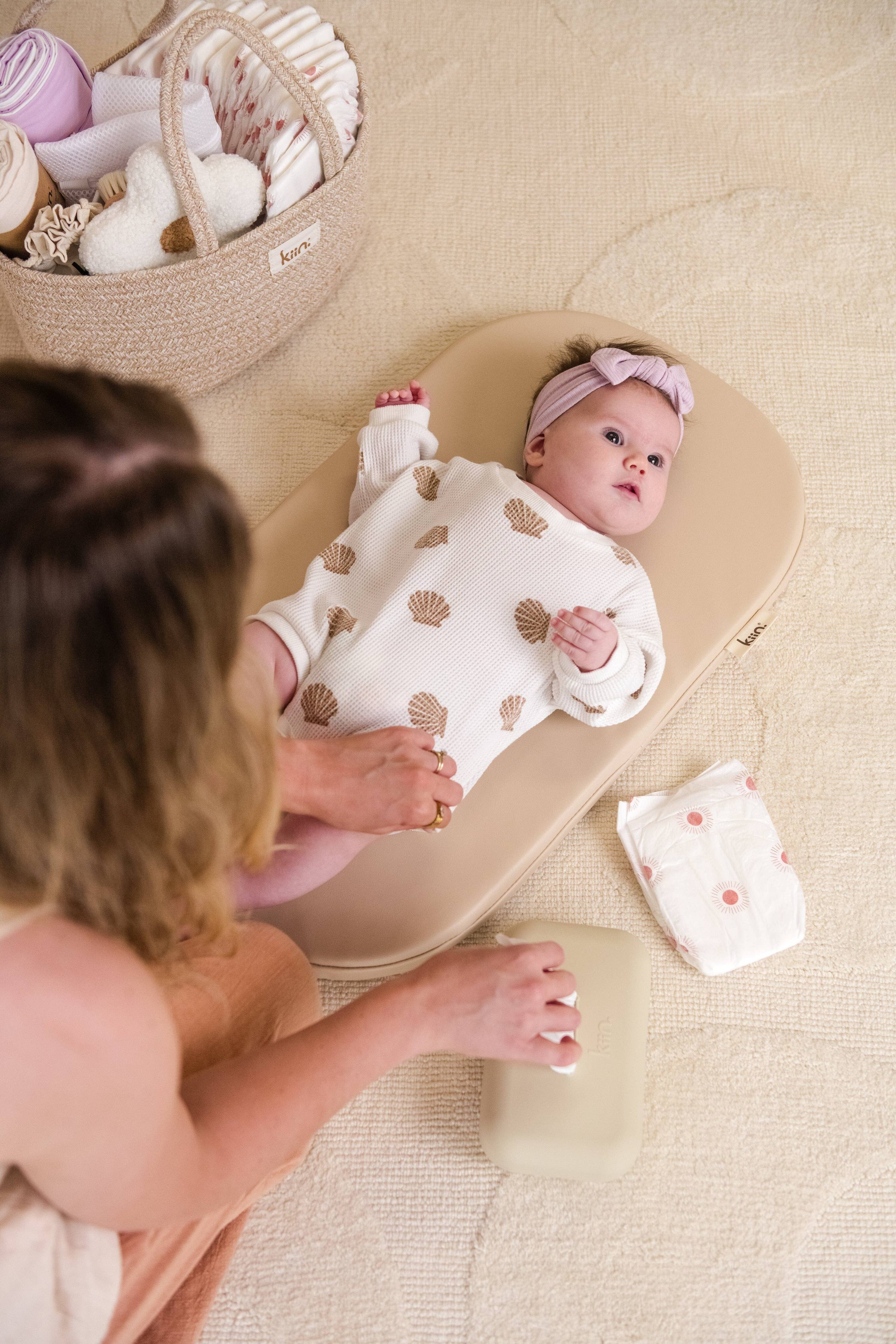 Baby lying on a wipeable change pad with a woman sitting nearby, surrounded by toys.