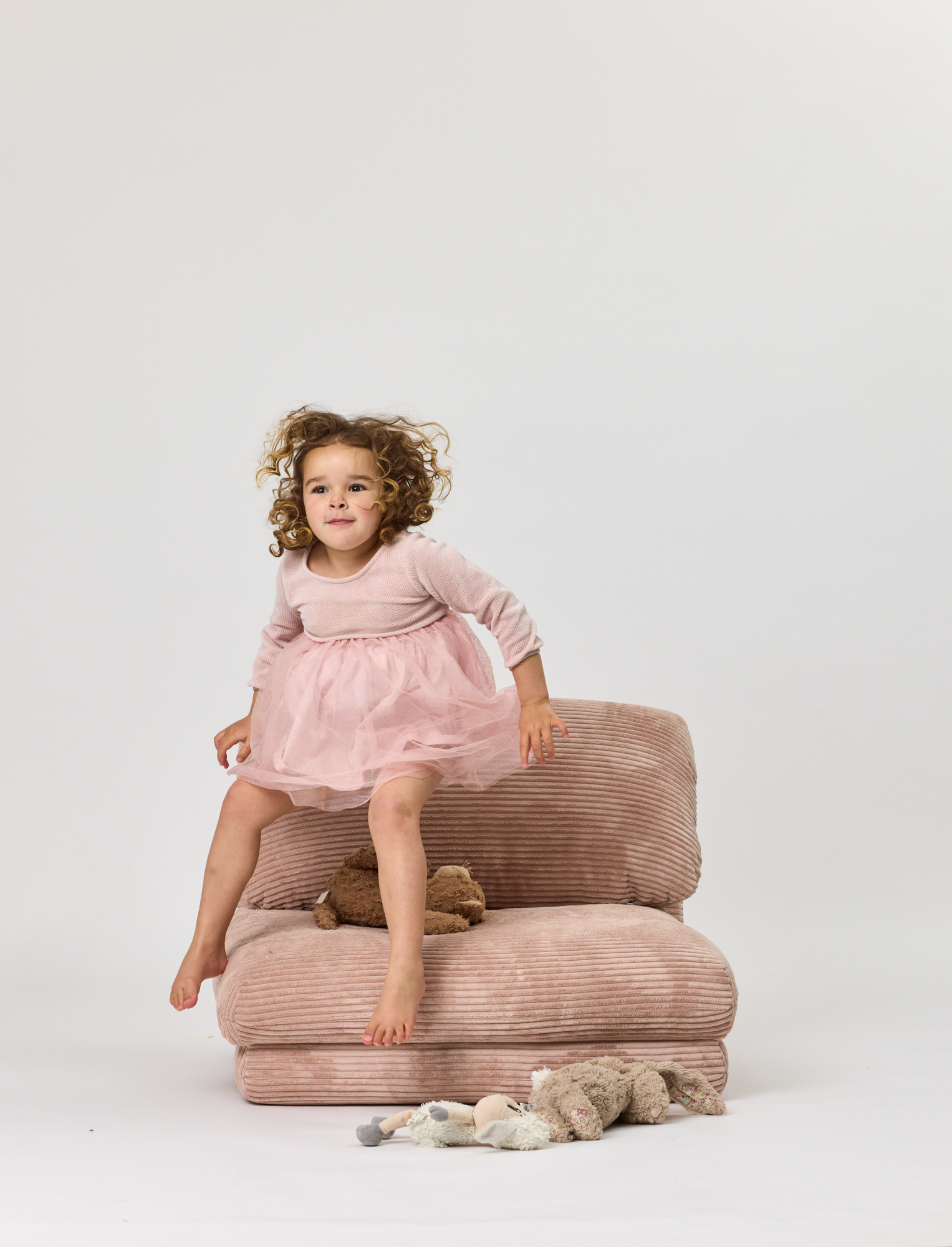 Child in a pink dress sitting on a plush chair with a white background