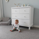 Baby sitting on the floor in front of a Florence dresser in a room with gray walls and carpet.