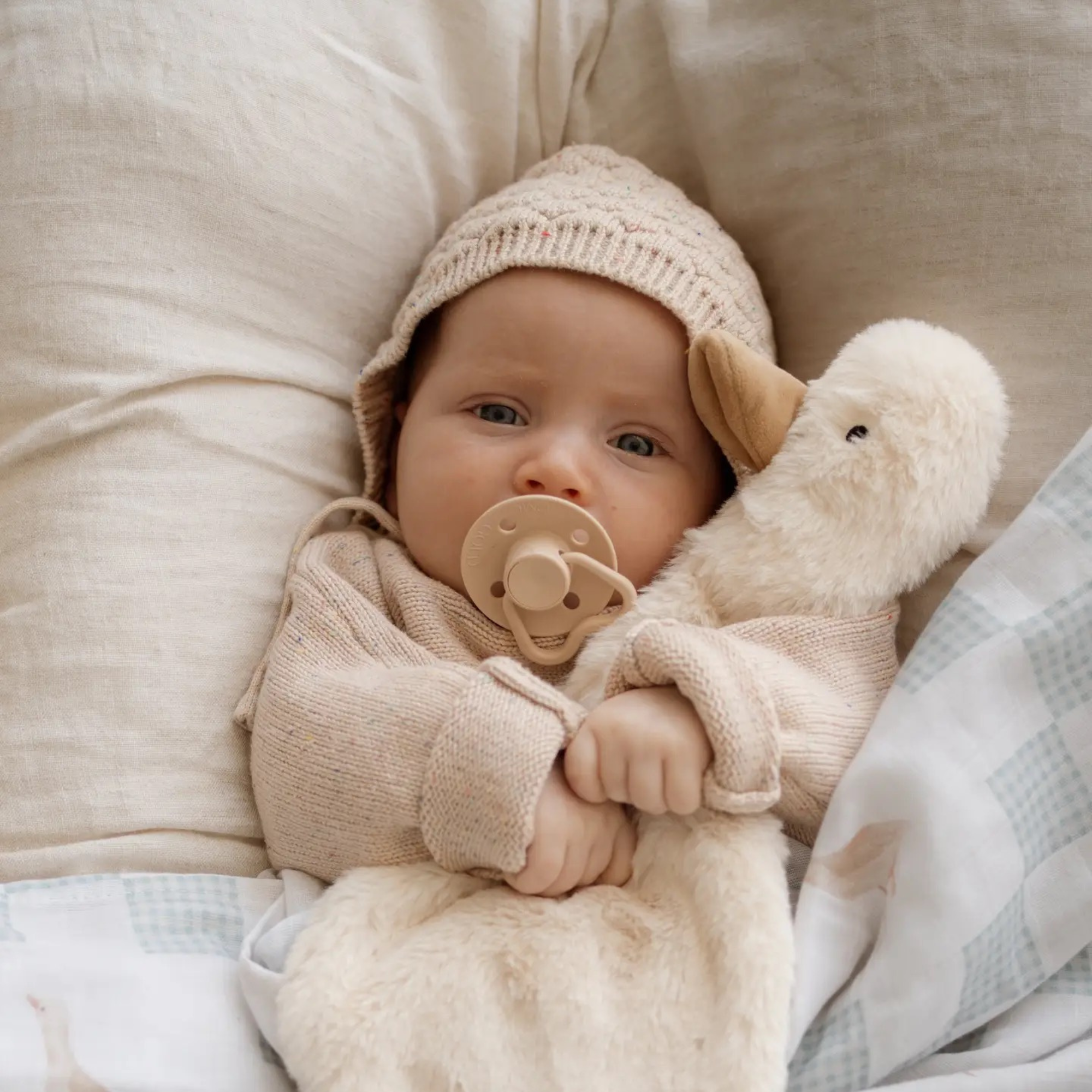 Baby in a beige outfit with a pacifier and Waddles the Goose Comforter Toy, lying on a soft surface.