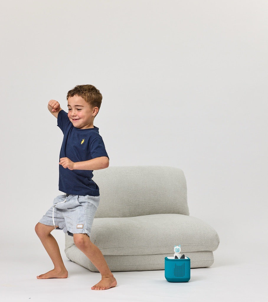 Child in navy shirt and light gray shorts standing next to a Rylee Recliner with a blue toy on a white background