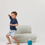 Child in navy shirt and light gray shorts standing next to a Rylee Recliner with a blue toy on a white background