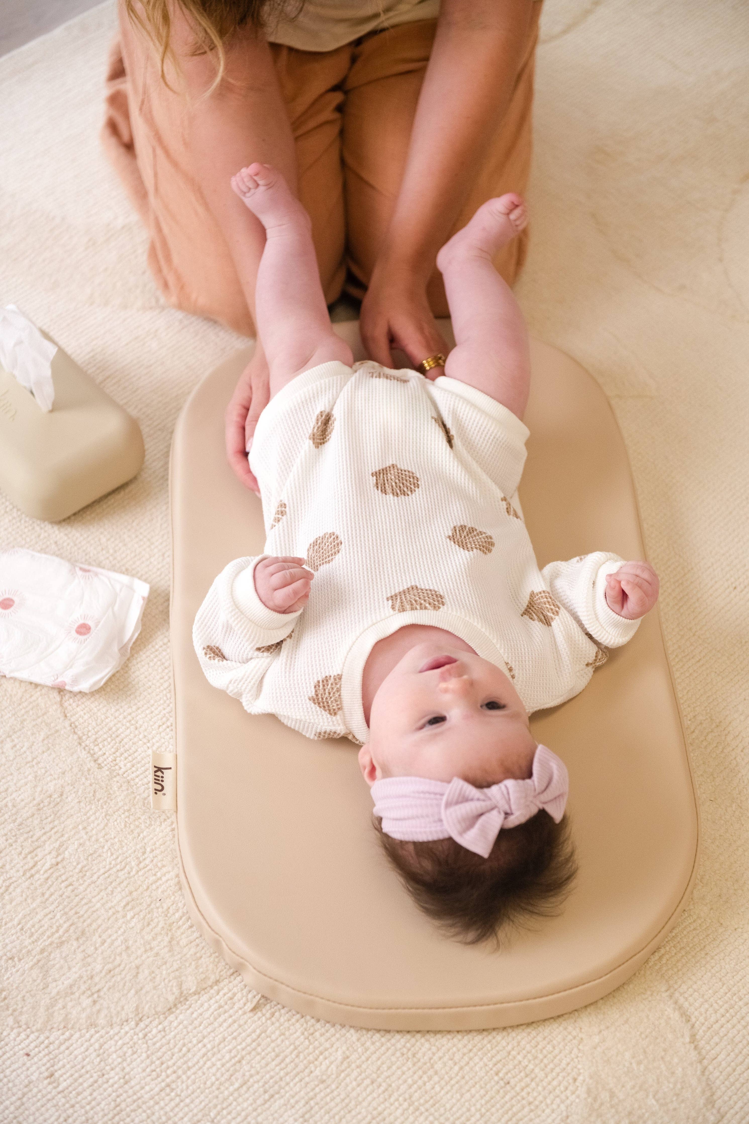 Baby lying on a wipeable change pad with a mother holding their feet, on a light-colored carpet.