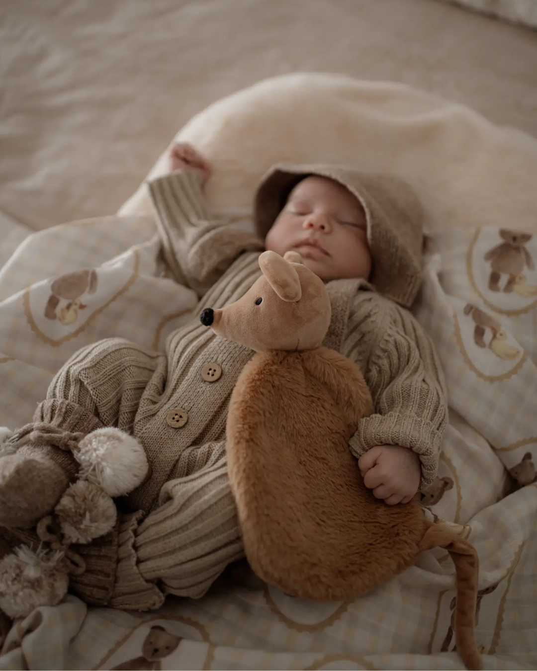 Baby in a knitted outfit holding a Squeaky the Sweet Comforter Toy, surrounded by soft bedding.