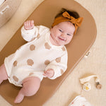 Baby lying on a wipeable change pad with a white outfit and orange headband, surrounded by baby items.