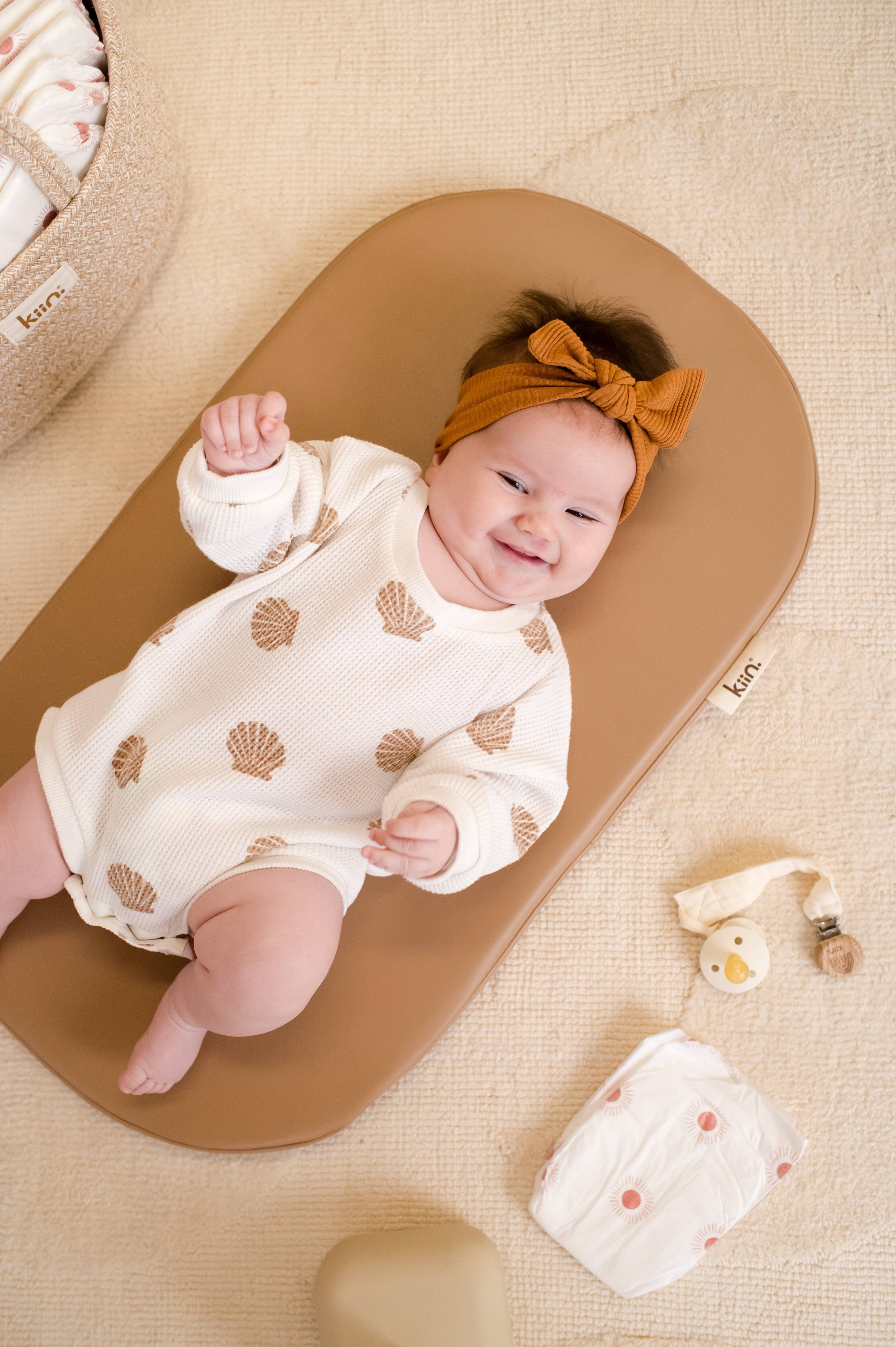 Baby lying on a wipeable change pad with a white outfit and orange headband, surrounded by baby items.